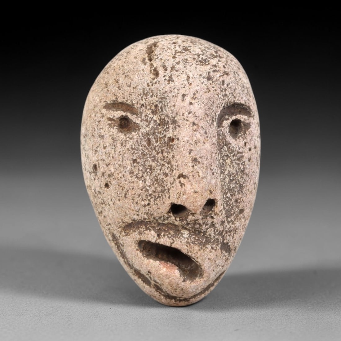 Ancient Stone Maskette - Medieval Figurine, 13FY: Carved from a teardrop-shaped stone pebble, this maskette features a stylized human face with incised details defining the eyes, nasal perforations, mustache, and open mouth. Stylistically consistent