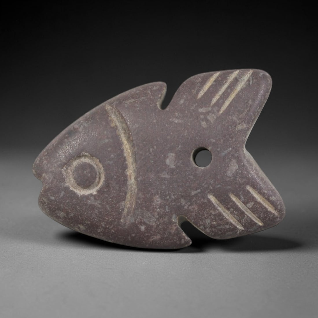 Ancient Stone Fish Amulet - Egyptian Animal Figurine, 52AP: Carved from a dense, muted purple stone with natural mineral inclusions and a weathered patina, this amulet depicts a stylized fish form with a pointed head, clearly defined circular eye, and a body t