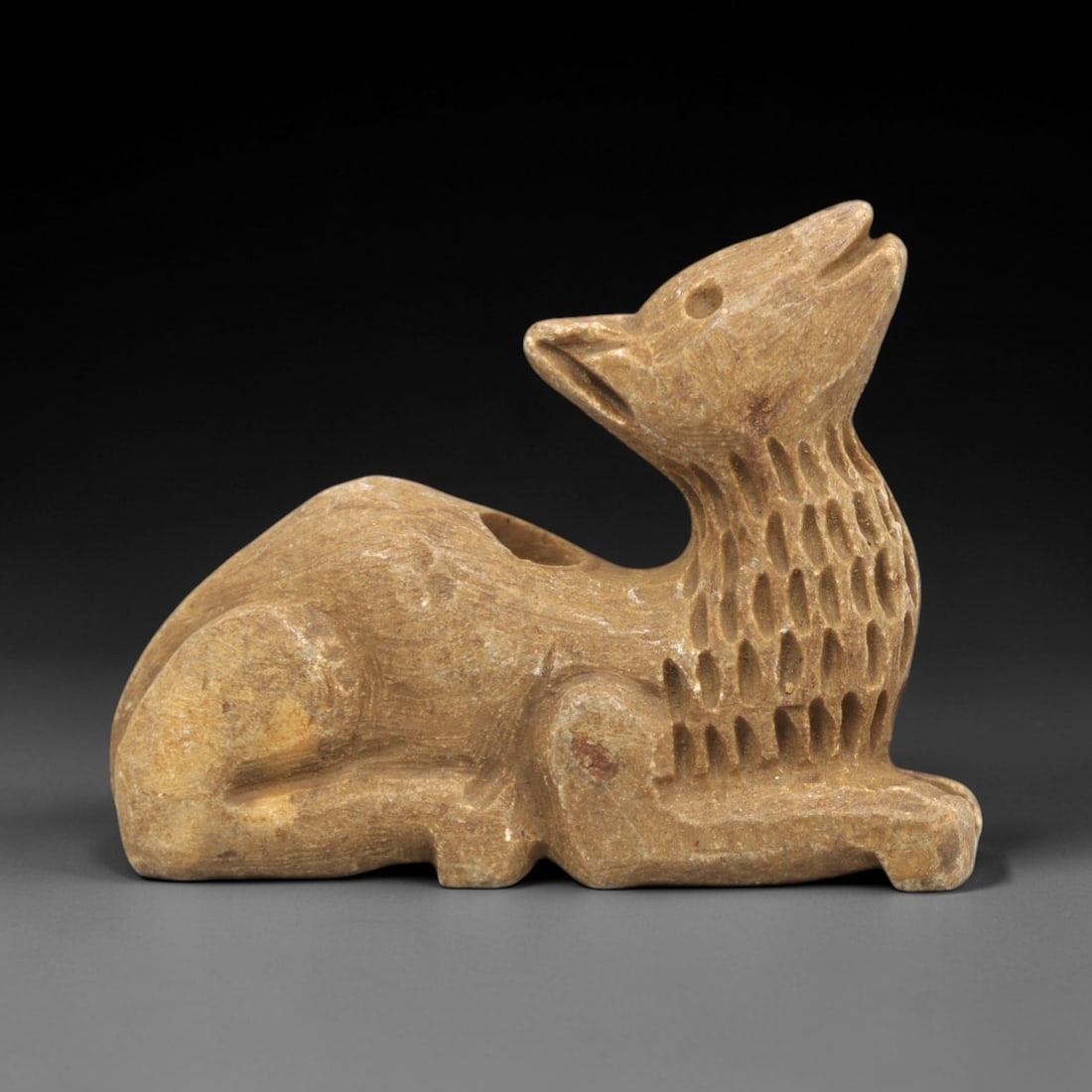 Ancient Stone Canine Figurine - Near Eastern Ritual Relics, 78ZP: Carved from warm tan stone, this reclining canine figurine features a stylized head tilted upward, with incised eyes, pointed ears, and an open mouth. The neck displays a textured, ribbed pattern, whi