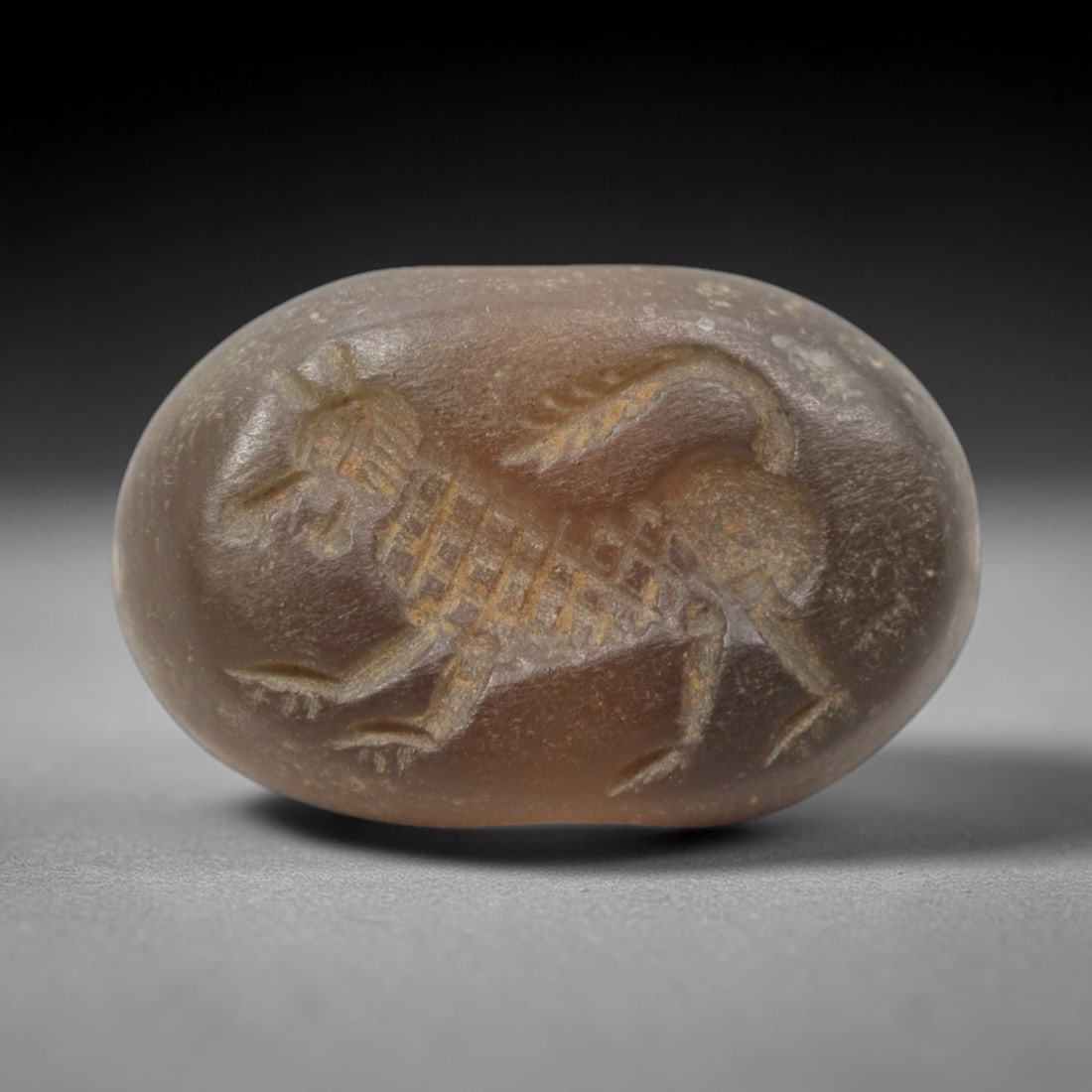 Ancient Chalcedony Stamp Seal - Near Eastern Beast Figure, 63ZS: Crafted from translucent chalcedony, this oval stamp seal bears an incised profile of a striding mythical beast, rendered with linear detailing to define its grid-patterned body, clawed limbs, and cur