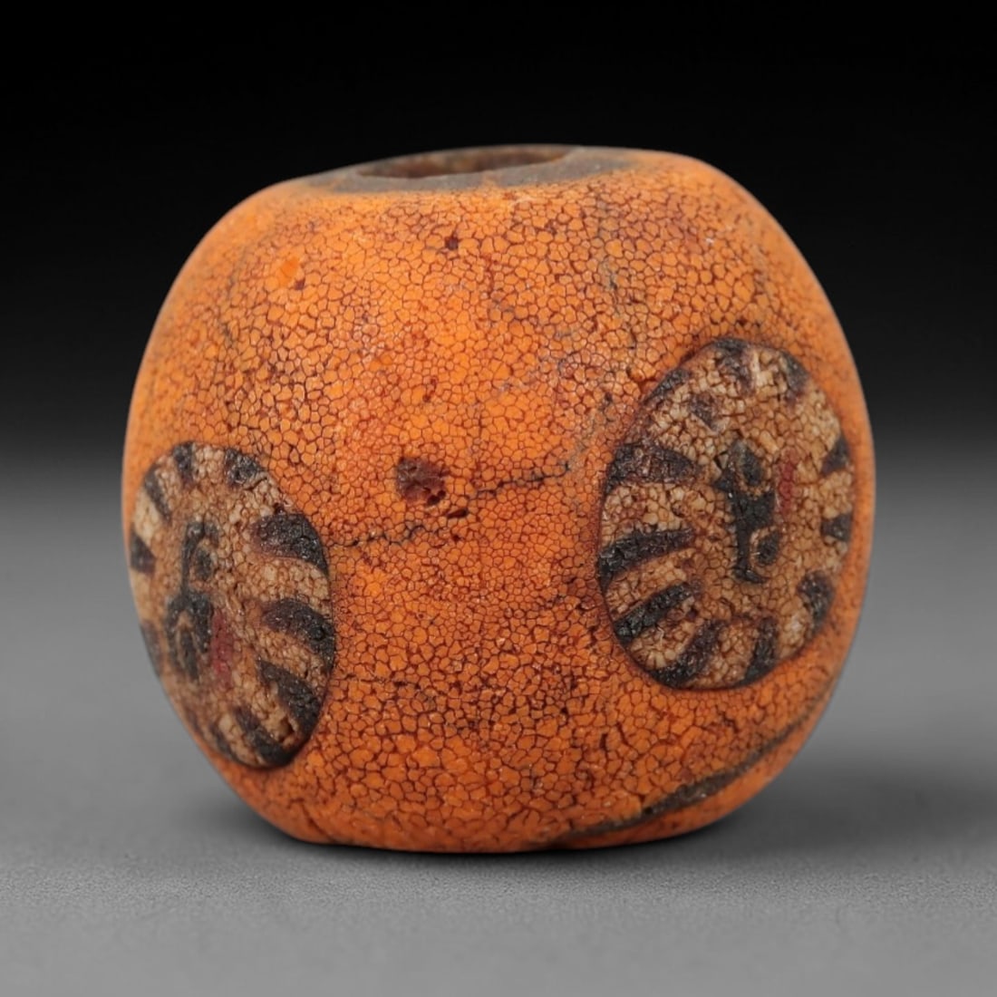 Ancient Glass Face Bead - Roman/Egyptian Mosaic Amulet Bead, 20FW: This spherical bead is crafted from porous orange glass, featuring two prominent circular medallions with stylized face motifs in black and white, evoking the protective face amulet traditions of the