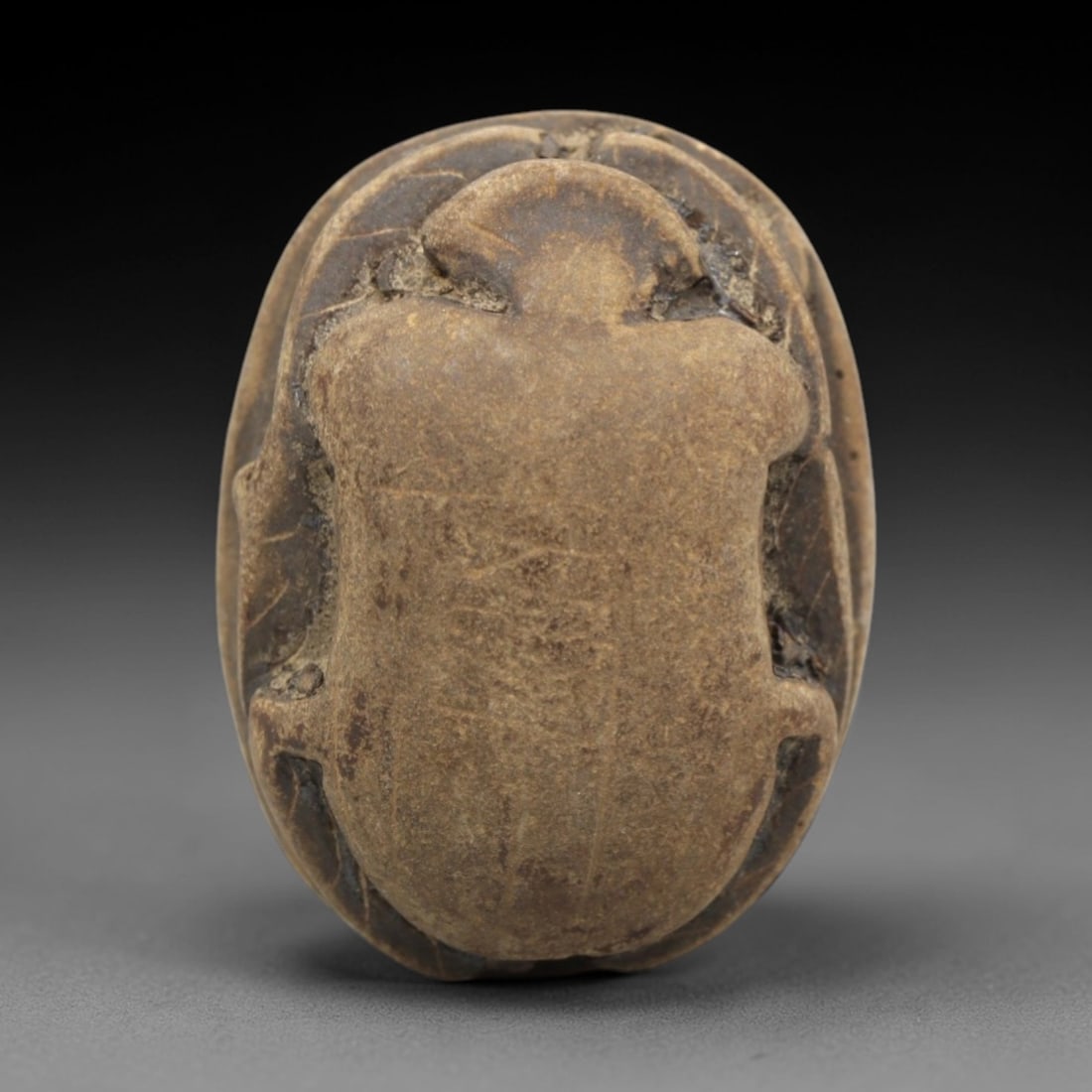 Ancient Egyptian Stone Scarab - Amulet Figurine, 52ZR: This artifact is a carved stone scarab amulet, fashioned from a dense, brownish-grey stone with a matte surface and subtle textural variations across its form. The dorsal side features a simplified ye