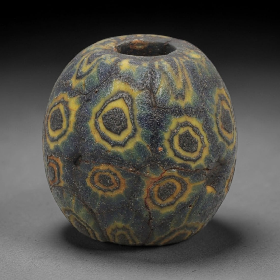 Ancient Glass Bead - Roman Egyptian Eye Amulet, 78ZN: Crafted from opaque glass with polychrome inlay, this ovoid bead features a central suspension hole and is decorated with repeating concentric eye motifs in yellow and dark grey glass against a dark m