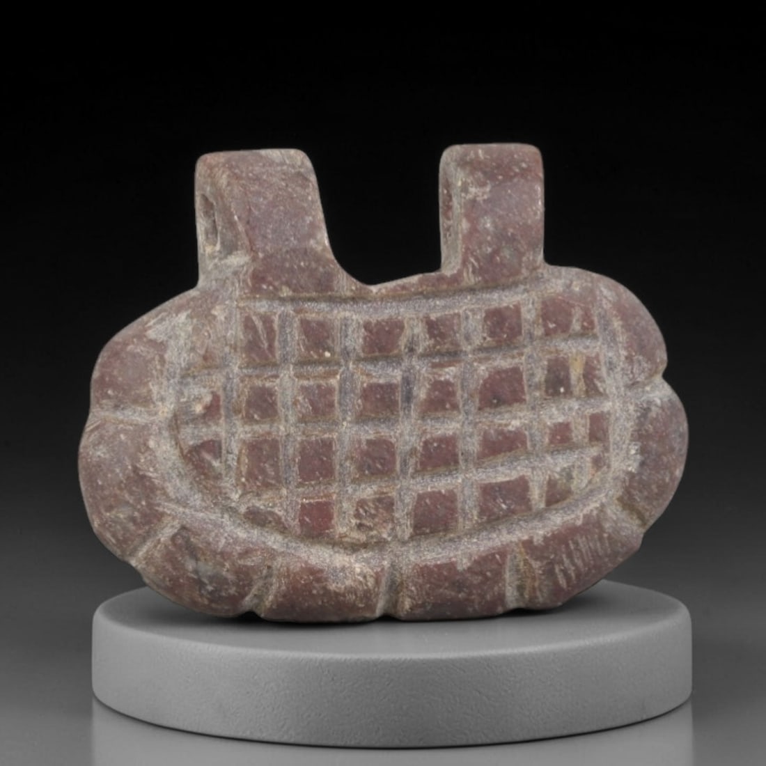 Ancient Stone Basket Amulet - Egyptian Ritual Figurine, 64AP: Carved from a dense, reddish stone with natural mineral inclusions and a weathered patina, this amulet takes the form of a stylized basket with a scalloped lower edge and two vertical suspension loops