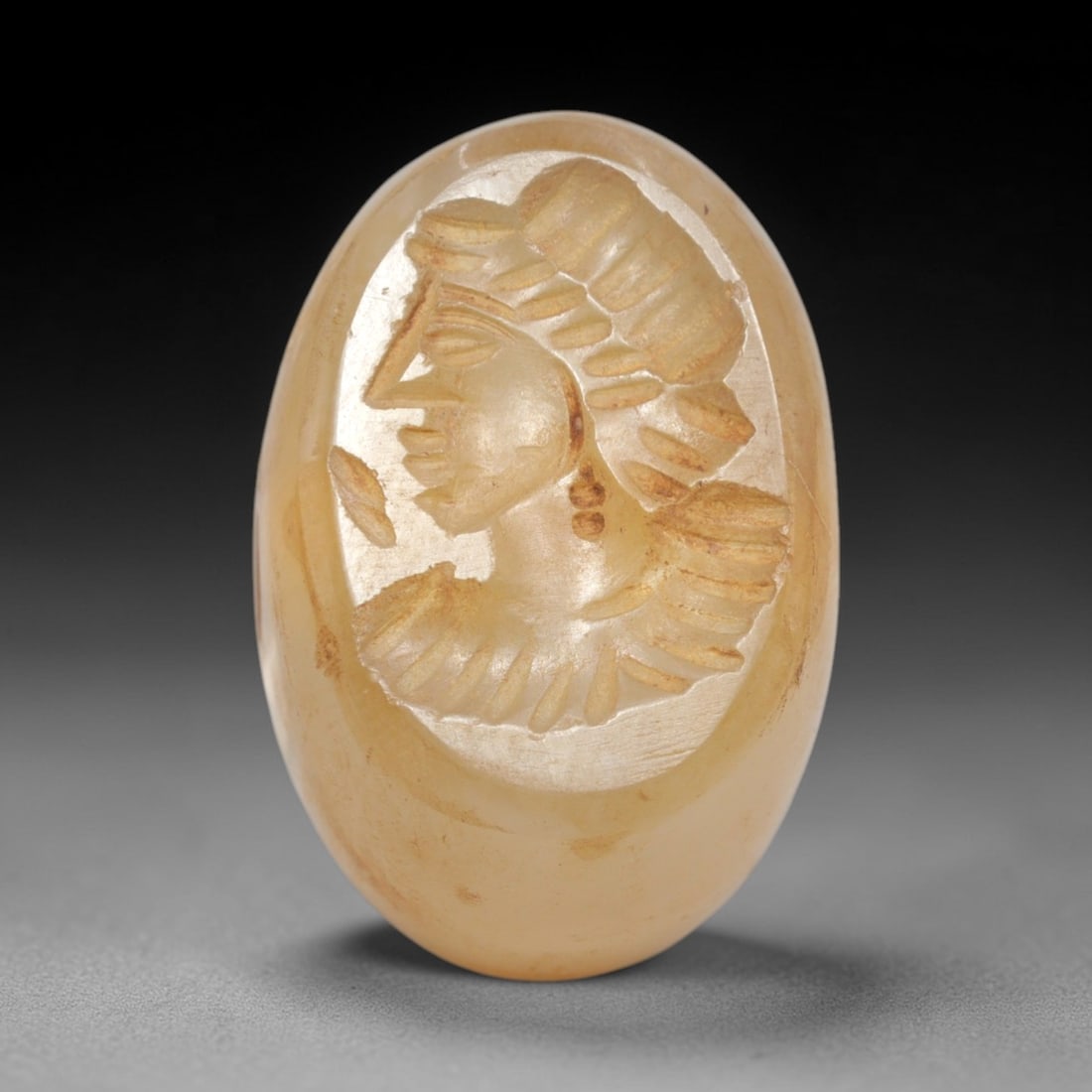 Ancient Chalcedony Stamp Seal - Near Eastern Female Bust, 67ZS: Crafted from translucent chalcedony, this oval stamp seal bears a deeply incised profile of a female bust, rendered with linear detailing to define her elaborate tiered headdress, facial features, and