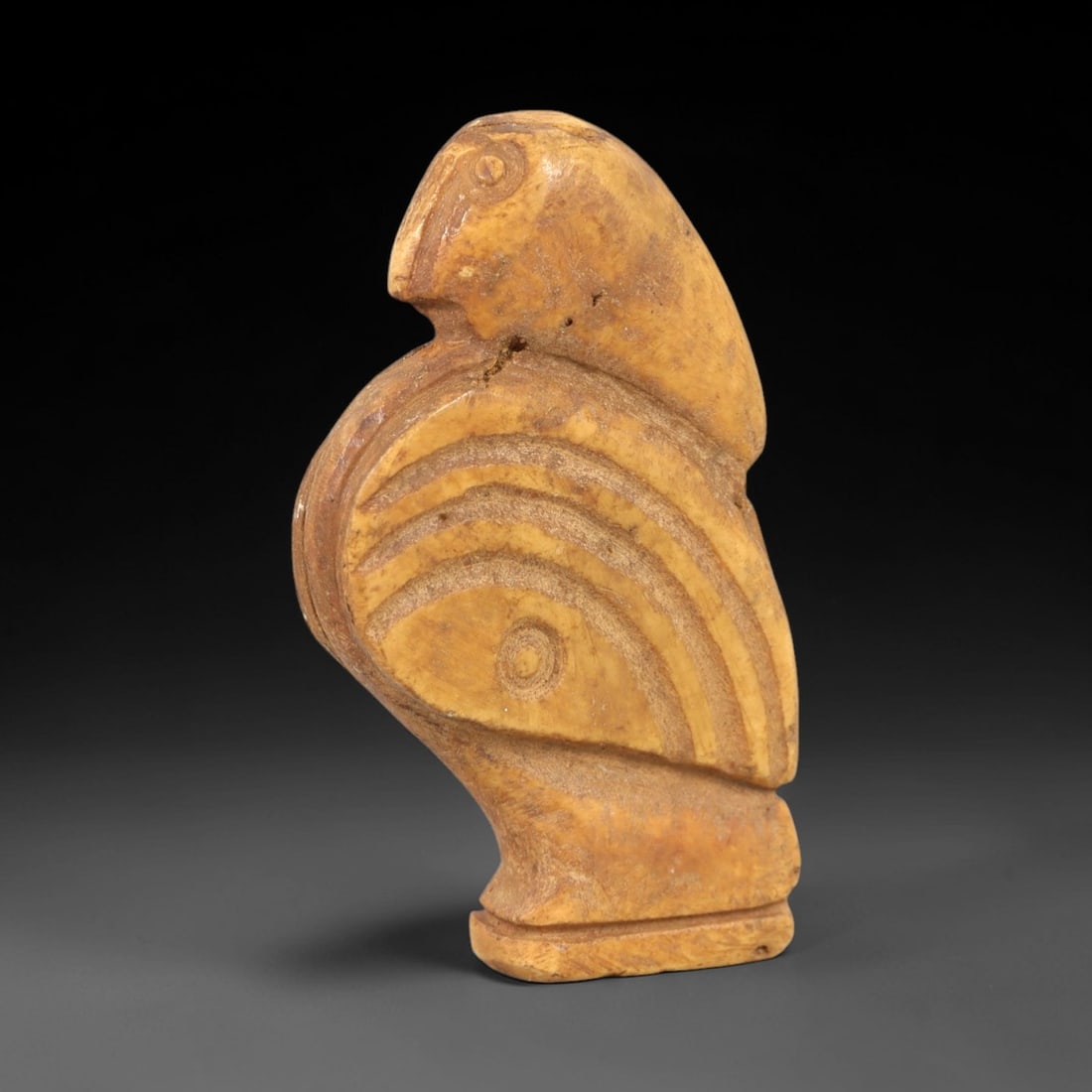 Ancient Bone Bird Amulet - Near Eastern Figurine, 84AL: Carved from tan bone, this amulet depicts a stylized avian form with a rounded head, incised eye, and curved beak. The body features concentric incised linear patterns creating a layered, feather-like
