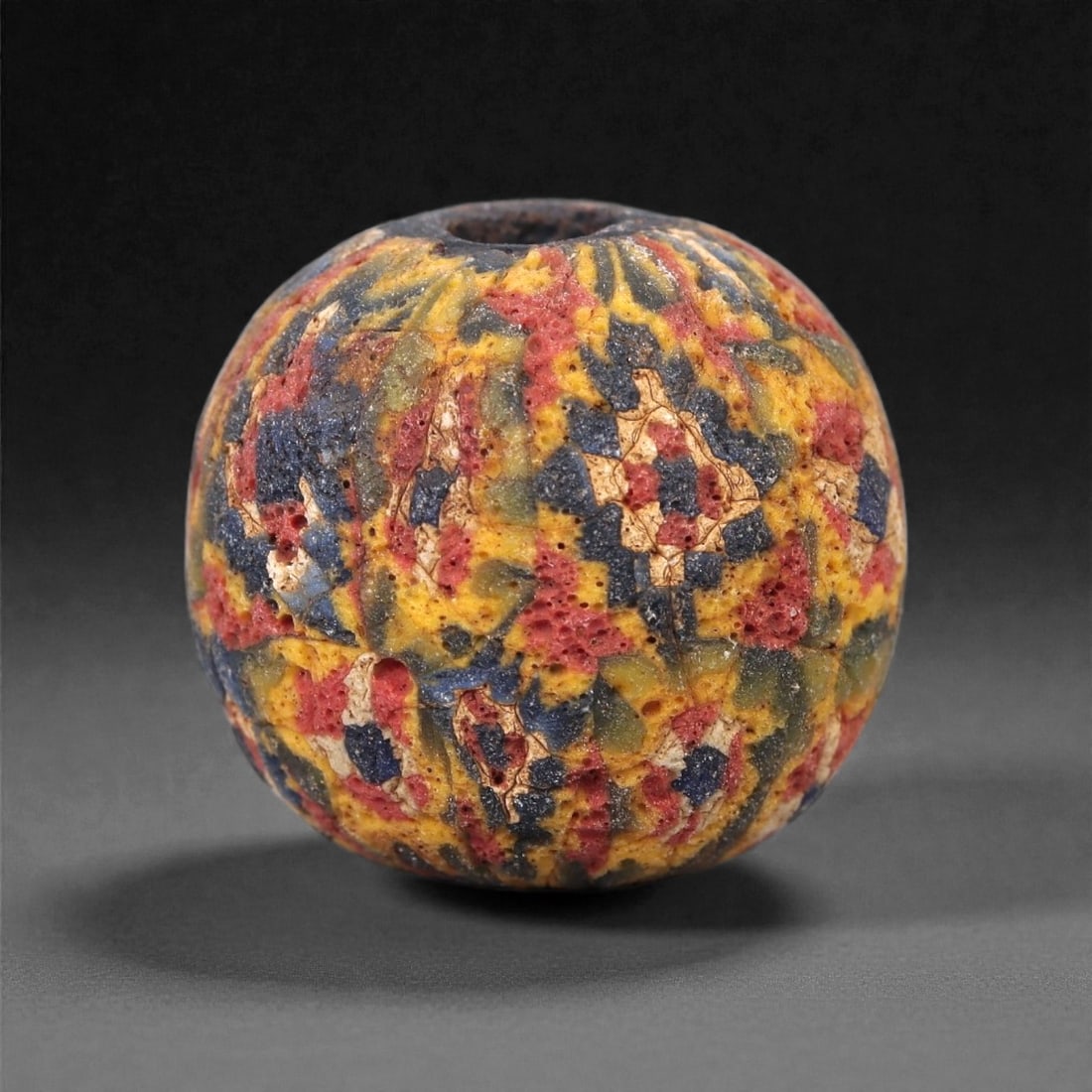 Ancient Glass Bead - Roman Mosaic Cane Sphere, 6Z: Formed of opaque and semi-opaque polychrome glass, this bead is produced using the mosaic cane technique, in which bundled glass rods were fused, sliced, and shaped to create a dense multicolored patt