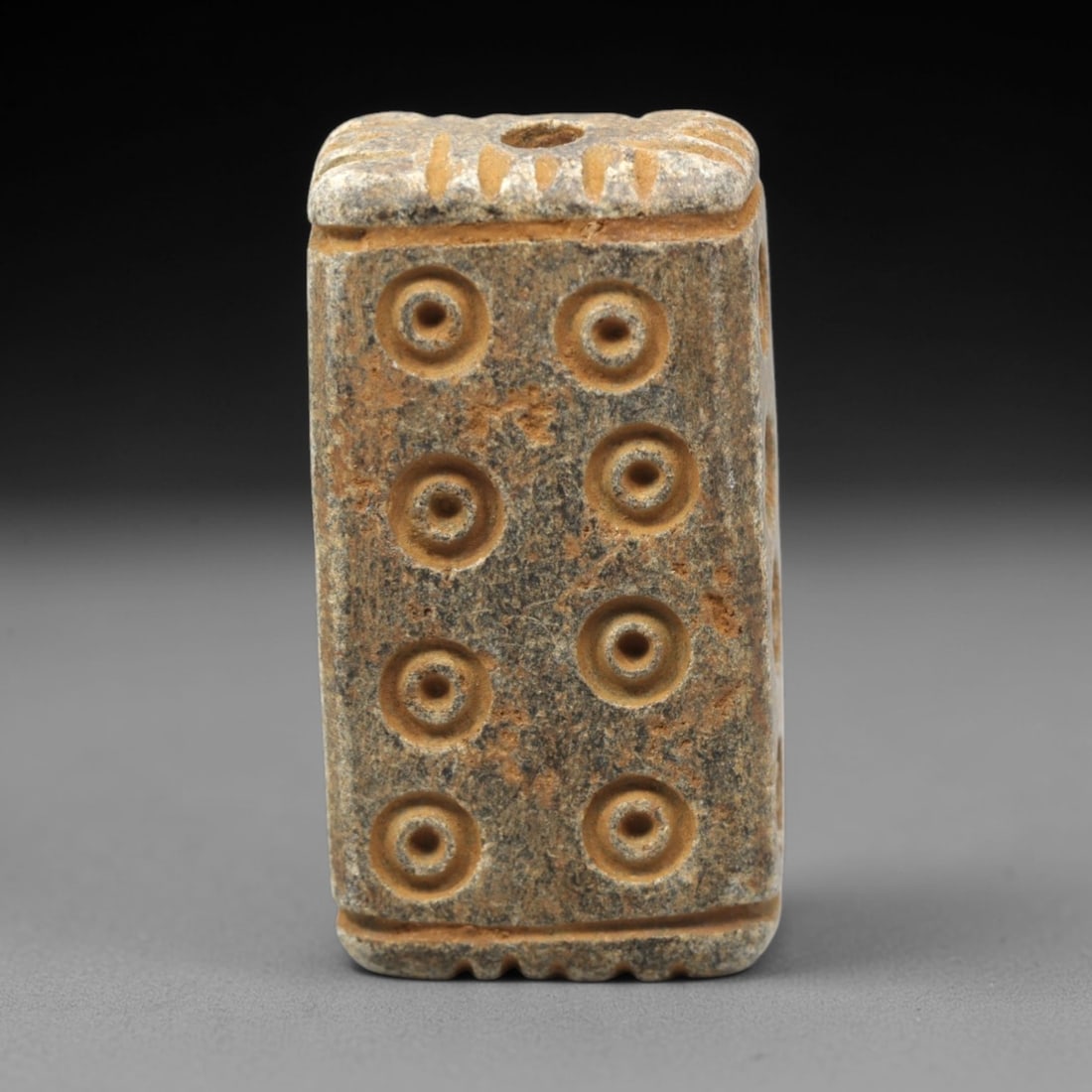 Ancient Steatite Amulet Bead - Near Eastern Concentric Circles, 50XV: Carved from dense steatite, this ancient amulet bead takes a rectangular form, with a vertical suspension hole at the top for wear. Its face is decorated with a symmetrical arrangement of eight concen