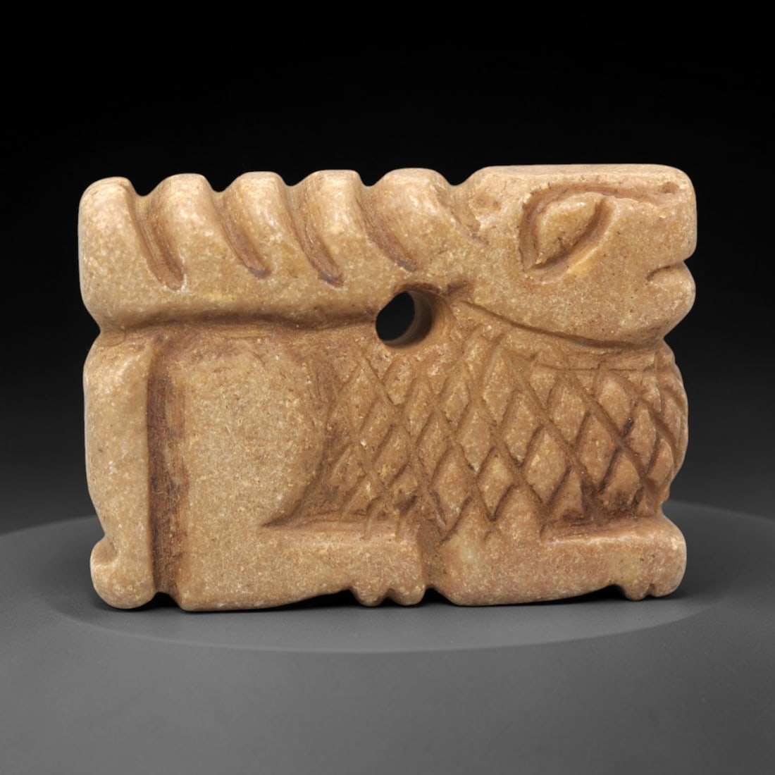 Ancient Steatite Caprid Amulet - Near Eastern Figurine, 48XV: Carved from steatite, this amulet depicts a stylized caprid (goat or sheep) in a crouched posture, with incised linear striations defining the curved horns and a diamond-patterned lattice across the t