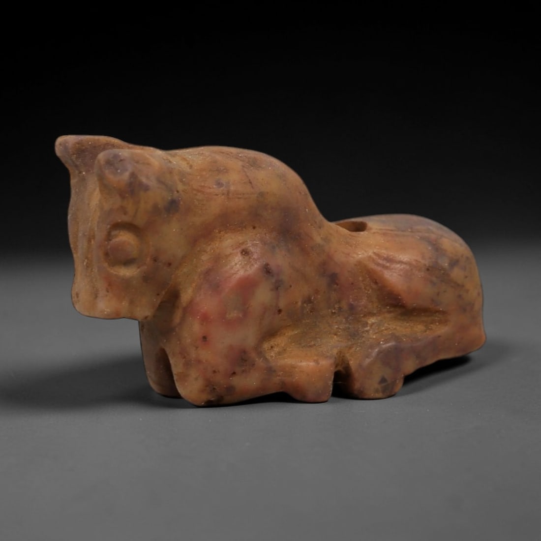 Ancient Mottled Stone Bovid Amulet - Near Eastern Figurine, 88AD: Carved from warm tan and reddish-purple mottled stone, this amulet depicts a stylized bovid in a compact, recumbent form. The creature features simplified facial details, incised circular eye motifs,