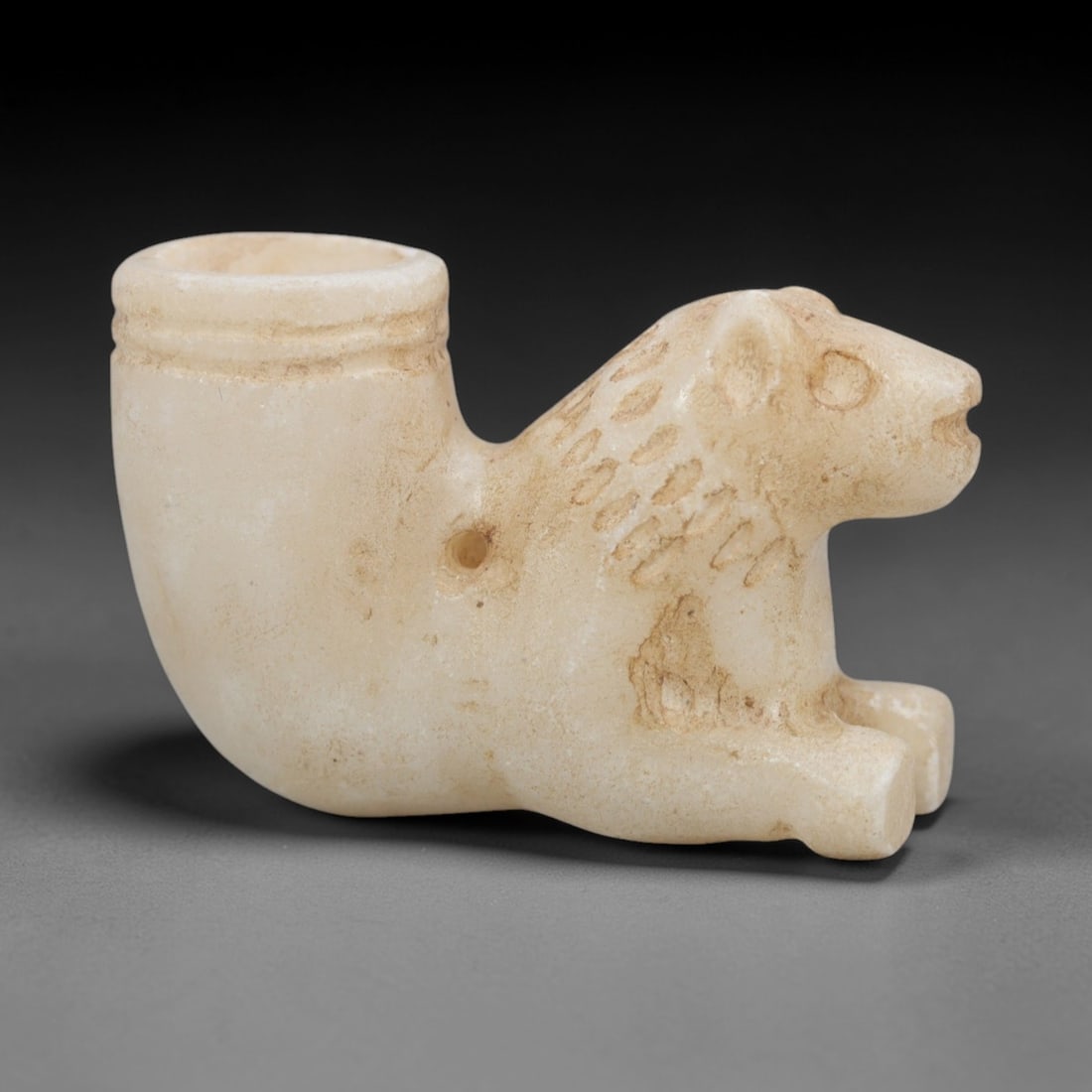 Ancient Marble Rhyton Amulet - Feline Form Figurine, 92XR: Crafted from pale marble, this rhyton-shaped amulet integrates a recumbent feline figure into its curved vessel form. The feline head, detailed with incised spots defining the pelt, rounded ears, and