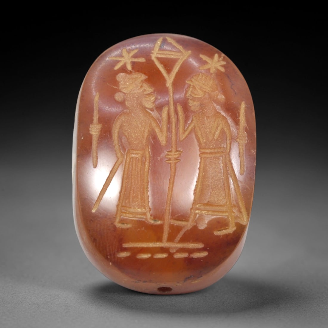Ancient Carnelian Intaglio - Near Eastern Figural Seal, 60ZP: This oval intaglio is carved from polished carnelian, featuring two standing male figures in relief, clad in pleated garments and head coverings, each holding a staff, flanking a central standard topp