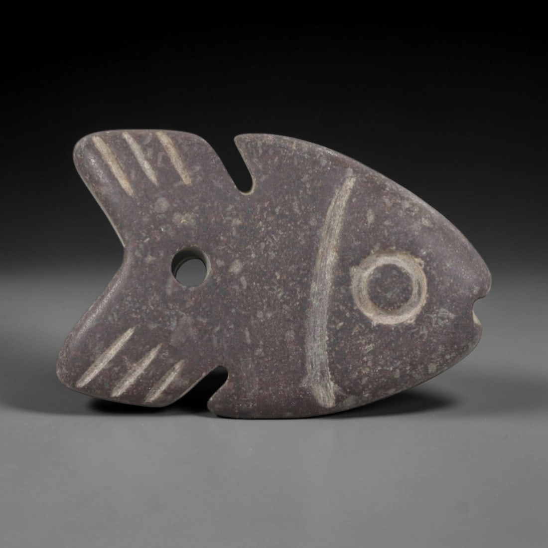 Ancient Stone Fish Amulet - Egyptian Ritual Pendant, 67AR: Carved from dark speckled stone, this fish amulet follows a stylized flat form with incised linear details defining the fins, tail, and facial structure, including a prominent circular eye motif. A pi