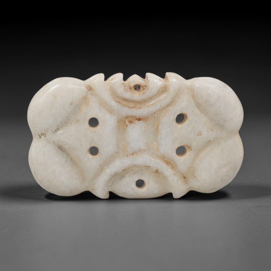 Ancient Marble Amulet - Near Eastern Ritual Pendant, 100XR: Crafted from pale marble, this amulet features a symmetrical, lobed form with curved, scalloped edges. The surface is punctuated by five circular suspension holes: one at the upper center, two on each