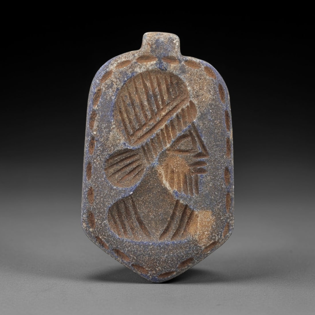 Ancient Lapis Lazuli Amulet - Near Eastern Profile Relief, 16FX: Carved from lapis lazuli, this shield-shaped amulet features a low-relief profile of a bearded male figure, with incised linear details defining the hairstyle, facial features, and upper body, framed
