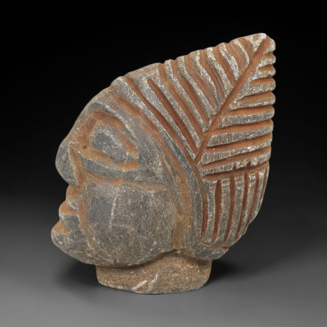 Ancient Stone Bust Amulet - Near Eastern Figurine, 29AM: This amulet is carved from hard stone, depicting a human bust in profile with stylized facial features and a prominent feathered headdress rendered in incised relief. The form reflects the iconographi