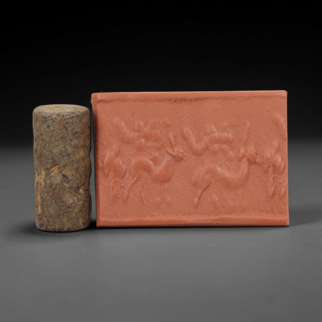 Ancient Near Eastern Cylinder Seal Amulet with Ibex Scene - Mesopotamian Figurine, 83AJ: Carved from speckled hardstone (likely serpentine or chlorite schist), this compact cylinder seal amulet features a finely incised pastoral scene of two leaping ibexes beneath stylized vegetal or cele