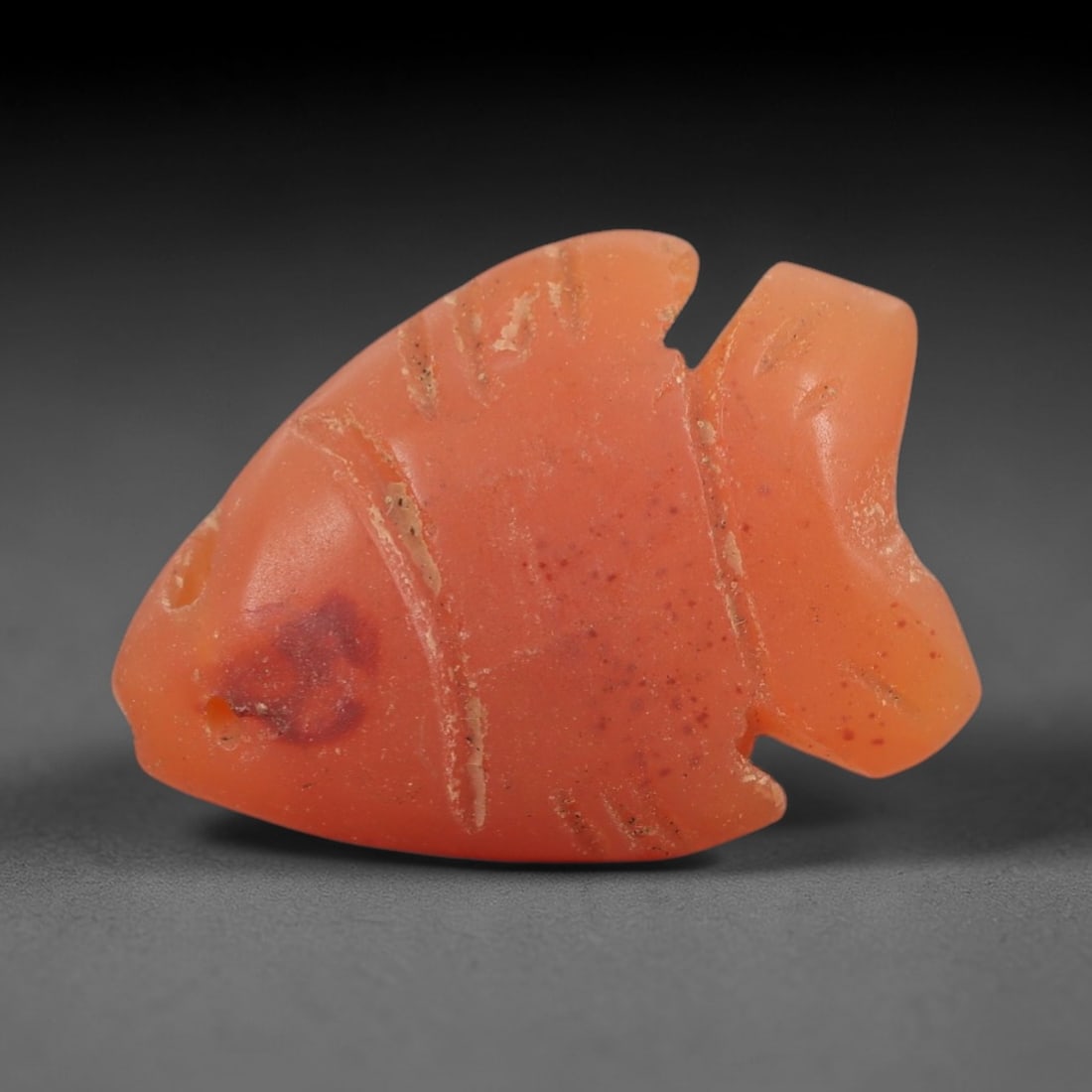 Ancient Carnelian Fish Amulet - Egyptian Figurine, 61AJ: Carved from translucent orange carnelian, this amulet is formed as a stylized fish with incised linear markings denoting fins and body segments, and a visible suspension hole at the head for wear. The