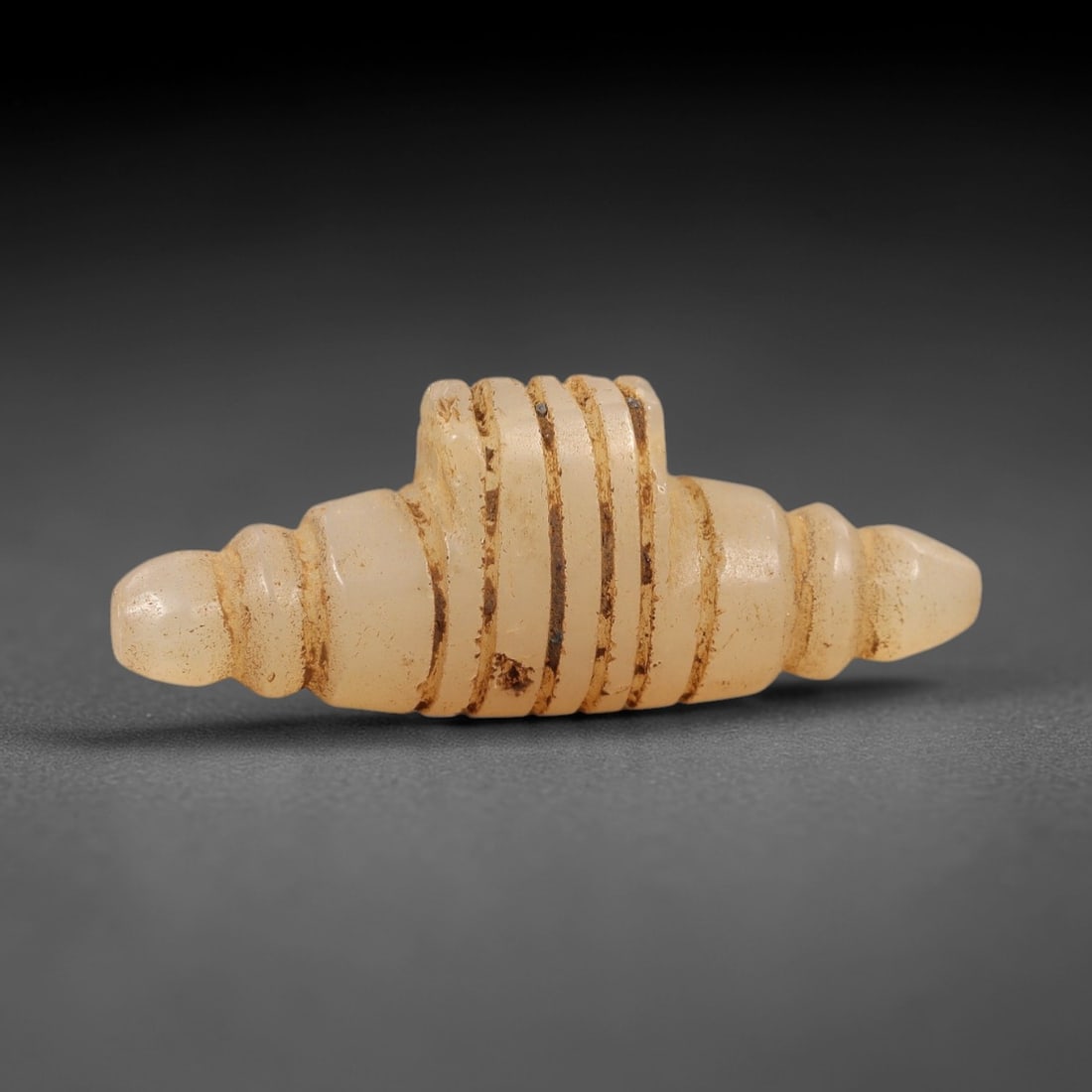 Ancient Chalcedony Amulet - Achaemenid Ritual Figurine, 83AL: Carved from translucent pale chalcedony, this Achaemenid amulet features a symmetrical, segmented form with parallel incised grooves encircling its central body, flanked by tapered, rounded ends. The