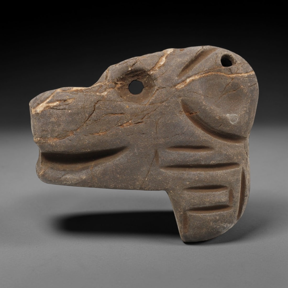 Ancient Stone Canine - Tribal Animal Amulet, 39FX: Carved from a compact block of grey-brown stone, this piece depicts a stylized canine head in profile, featuring a pronounced snout, open mouth, and incised linear details suggesting facial structure
