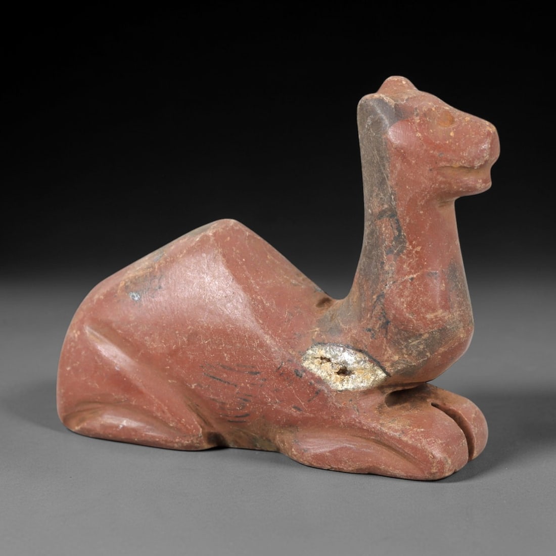 Ancient Stone Camel Effigy - Near Eastern Animal Figurine, 47AM: This camel effigy is carved from solid reddish-brown stone with natural grey veining and mineral deposits, depicting a stylized camel in a recumbent, crouched posture. The piece features clean, incise