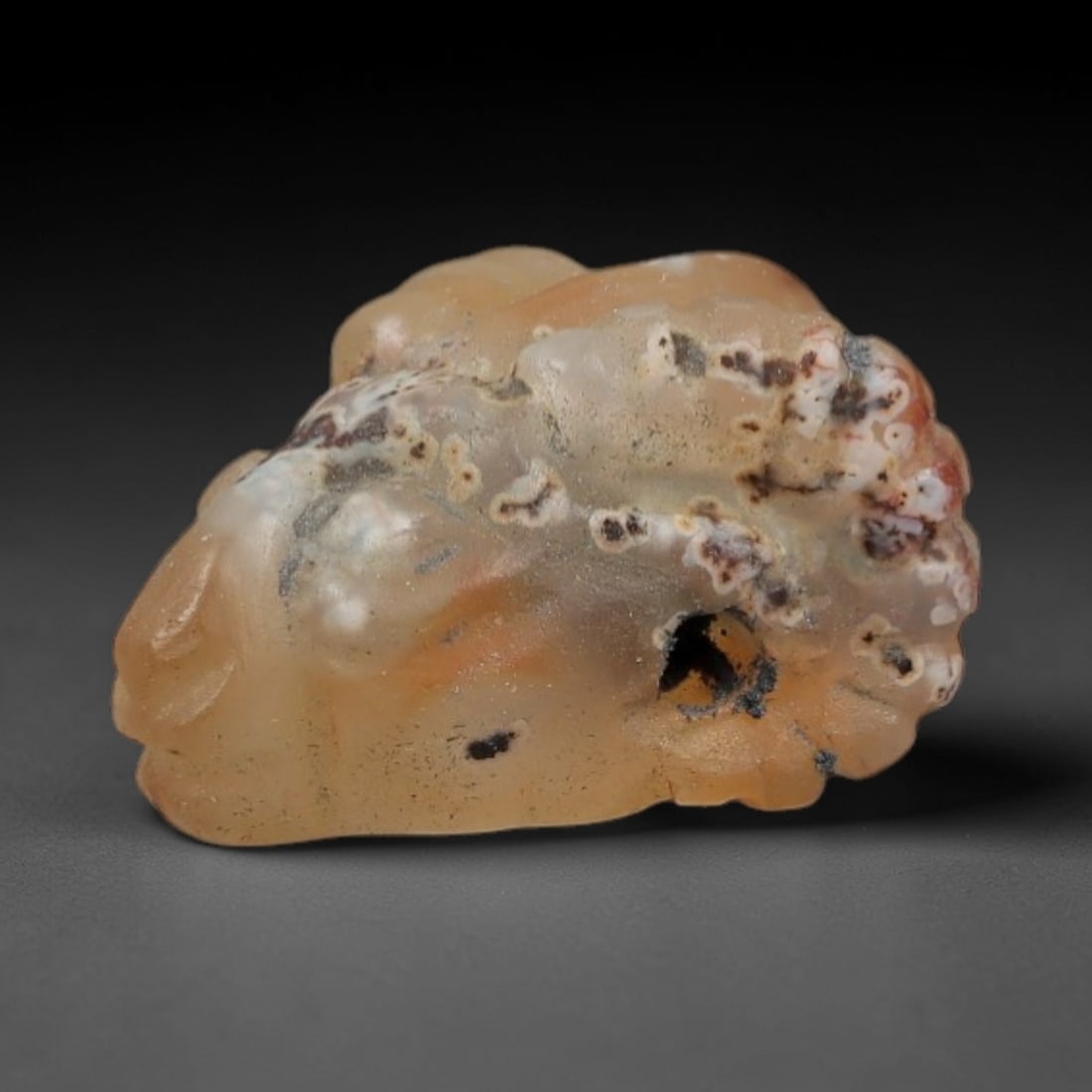 Ancient Agate Ram's Head Amulet - Near Eastern Figurine, 26AJ: Carved from warm, translucent peach agate with striking natural inclusions of deep red-brown, creamy white, and black dendritic patterns, this amulet is formed as a stylized ram's head with incised li
