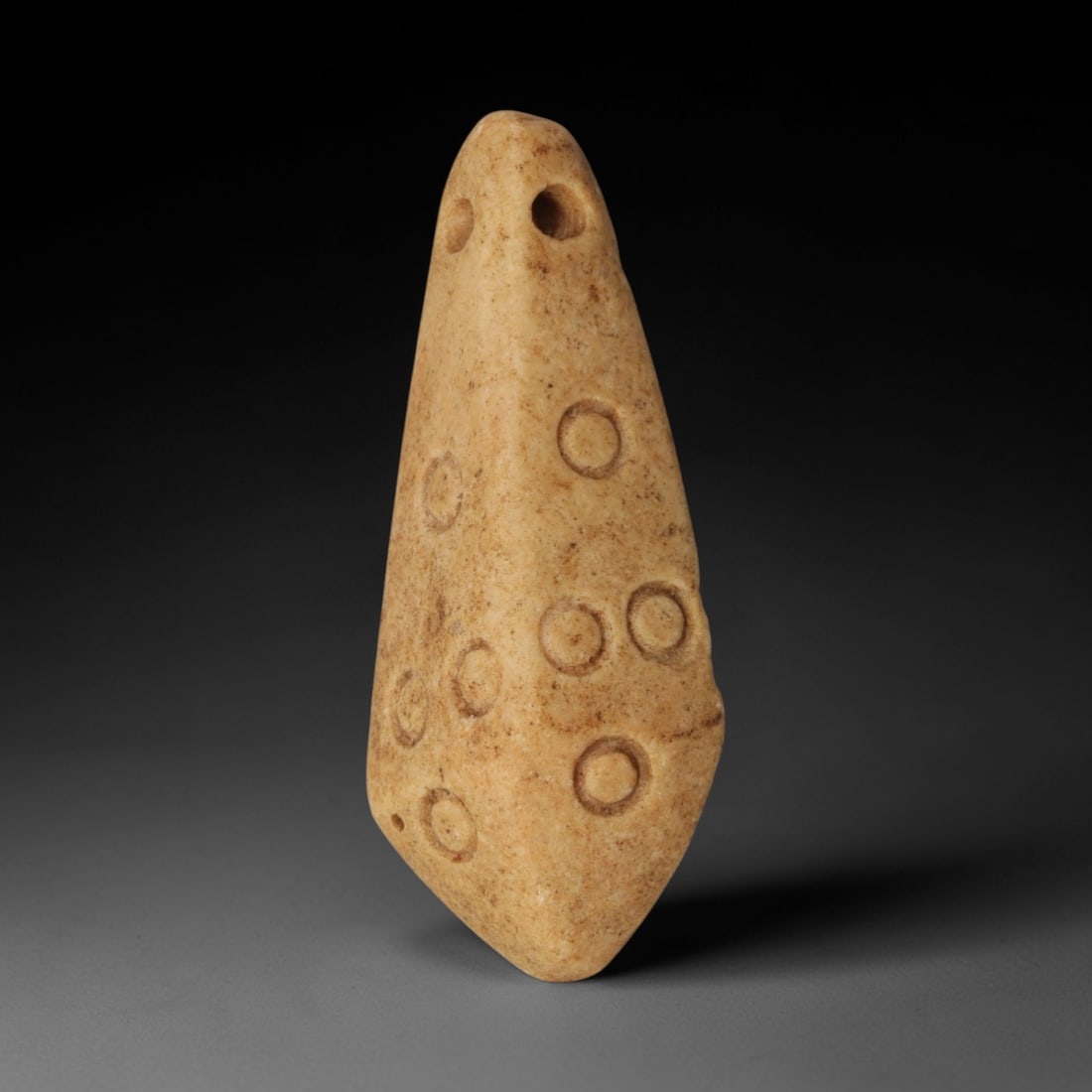 Ancient Stone Amulet - Near Eastern Circle-Carved Pendant, 56XP: Carved from fine-grained, pale tan stone, this pendant amulet features a tapering, pointed form with two suspension holes at the apex, designed for wearable ritual use. Its surface is adorned with inc