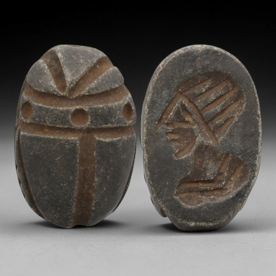 Ancient Near Eastern Scarab Stamp Seal - Anthropomorphic Profile Intaglio, 26ZS: Carved from a dense, dark grey stone, this oval scarab stamp seal features a classic scaraboid form on its dorsal side, with incised linear patterns defining the beetle's elytra, central suture, and t