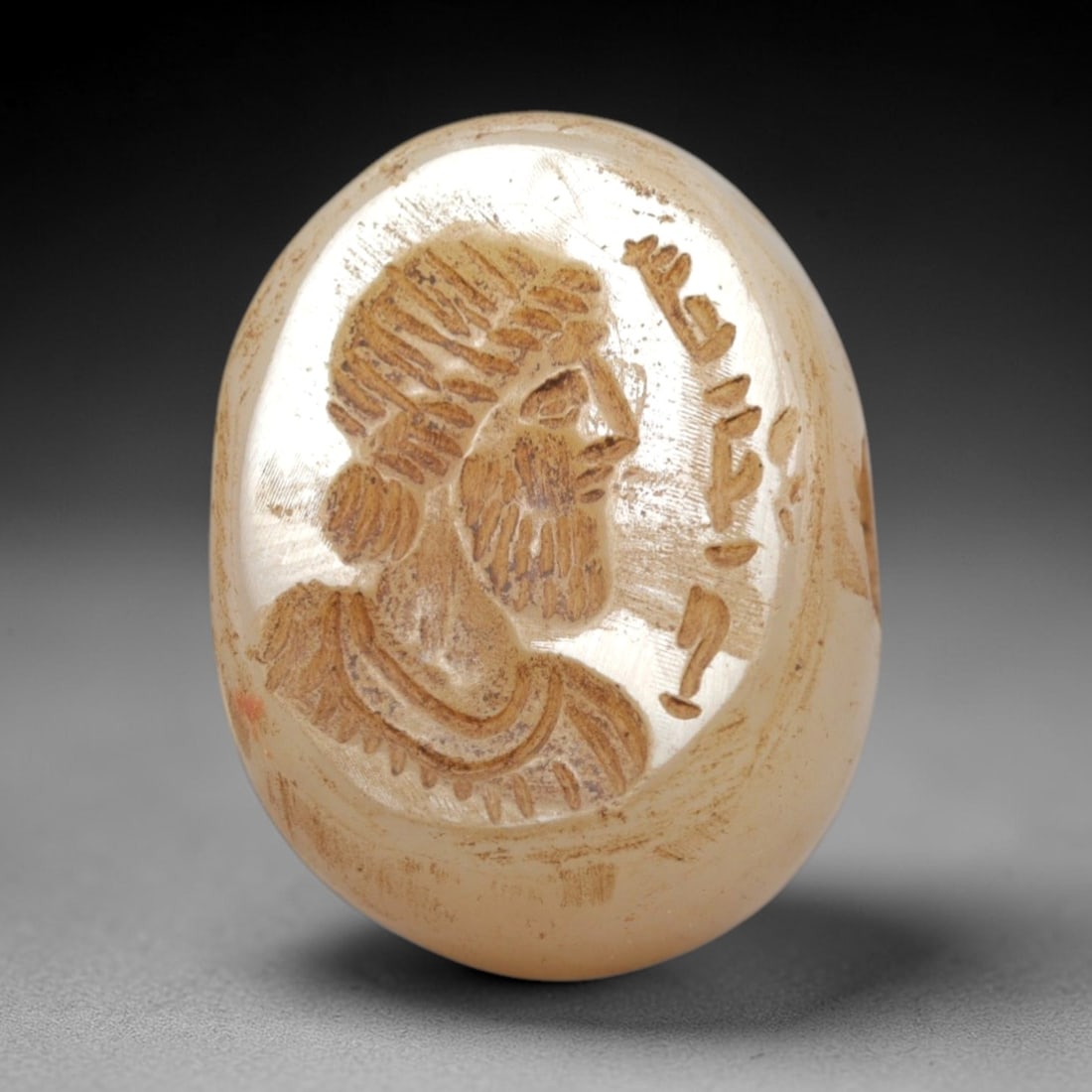 Ancient Chalcedony Stamp Seal - Near Eastern Bearded Figure, 54ZS: Crafted from translucent chalcedony, this oval stamp seal bears an incised profile of a bearded male figure, rendered with linear detailing to define his stylized headdress, full beard, and draped gar
