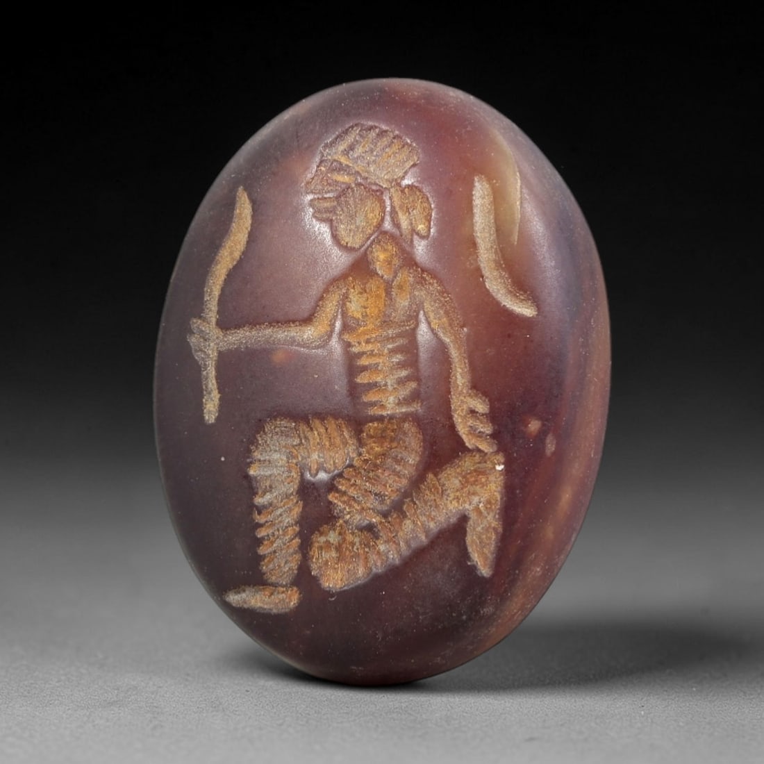 Ancient Agate Warrior Intaglio - Near Eastern Amulet, 31AR: This oval intaglio is carved from rich, deep-hued agate, featuring a high-relief depiction of a kneeling warrior figure in profile, holding a curved blade. The figure is rendered with incised linear d