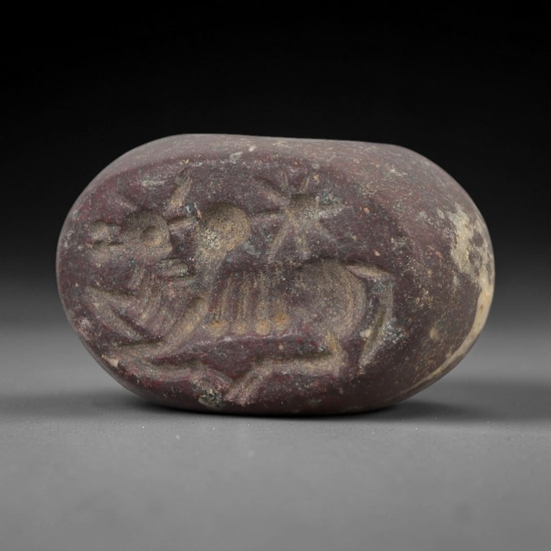 Ancient Red Stone Stamp Seal - Near Eastern Bovid, 83AN: Carved from dense, reddish-purple stone, this hemispherical stamp seal depicts a recumbent bovid in profile, characterized by prominent horns, a ribbed torso, and stylized limbs. A radiating sun-like
