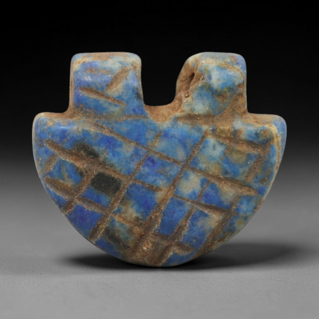 Ancient Lapis Lazuli Heart Amulet - Egyptian Ritual Pendant, 30ZT: Carved from lapis lazuli, this amulet is formed in a classic heart shape with two raised upper lugs framing a central suspension channel, its surface incised with a dense grid pattern of intersecting