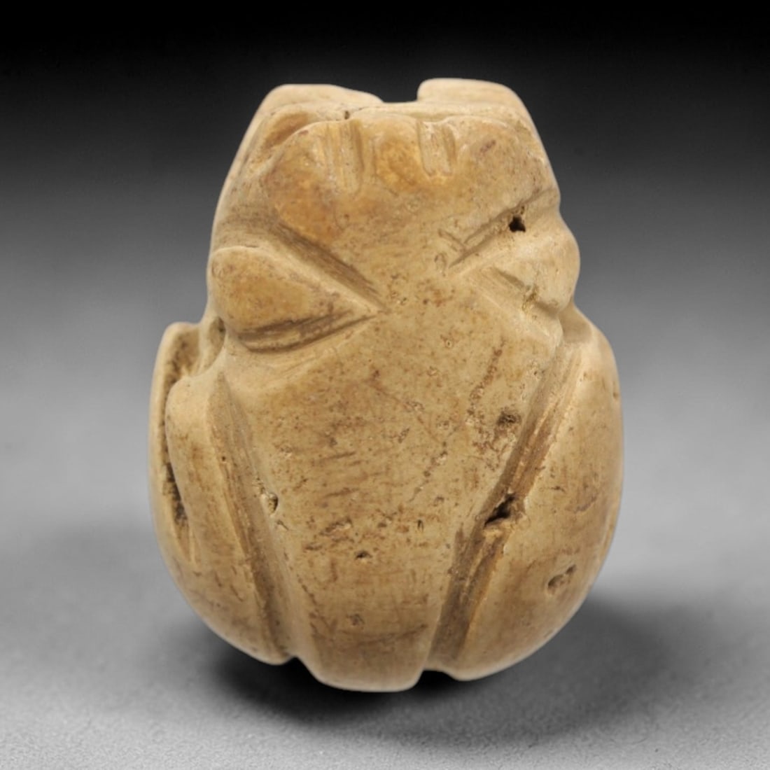 Ancient Limestone Scarab Amulet - Egyptian Ritual Figure, 64ZW: This amulet is carved from pale beige limestone, shaped in the classic form of a scarab beetle with incised details defining the head, elytra, and segmented body. A vertical perforation runs through
