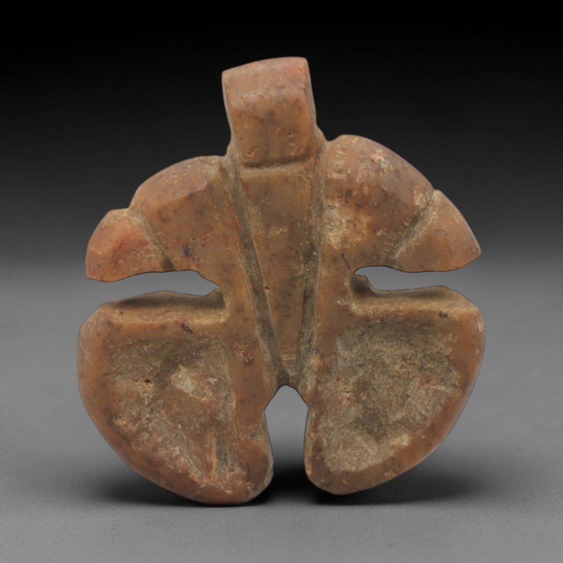 Ancient Stone Amulet - Near Eastern Double Axe Pendant, 5XR: This amulet is carved from a warm, reddish-brown stone, shaped into a symmetrical double axe form with a raised rectangular suspension loop at the top, confirming its use as a wearable protective char