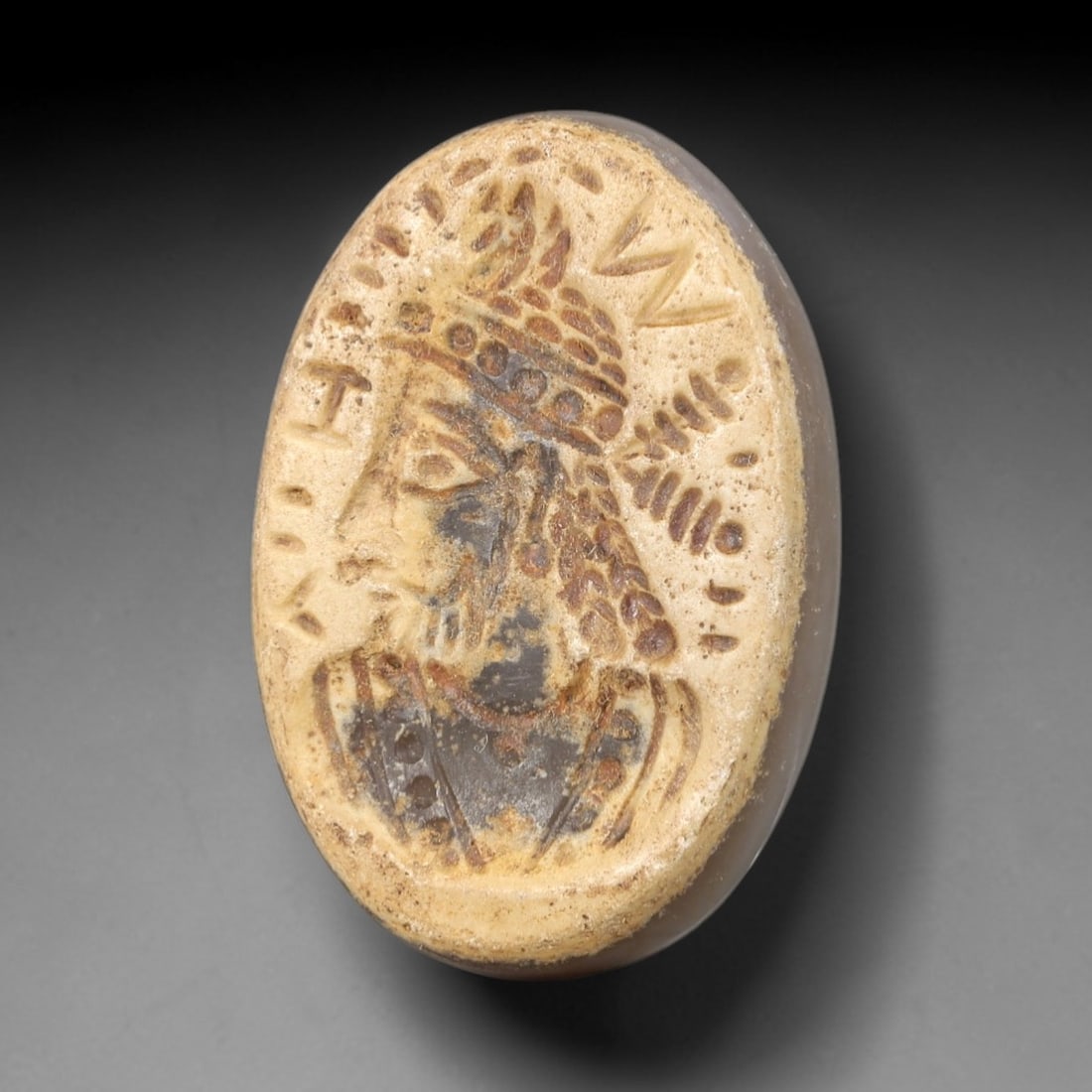 Ancient Stone Royal Bust Intaglio - Near Eastern Amulet, 69AR: This oval intaglio is carved from a layered stone matrix, featuring a high-relief profile depiction of a royal or divine figure, adorned with an elaborate beaded headdress and incised linear details d