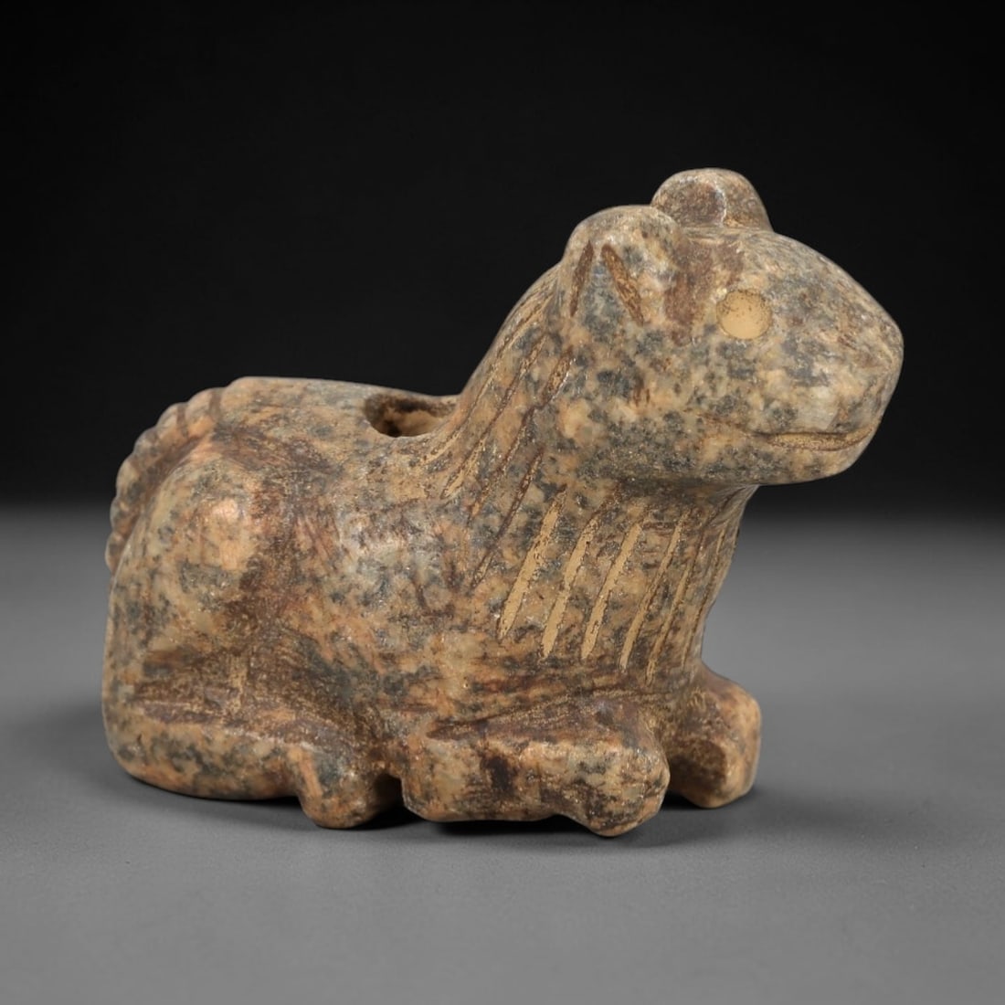 Ancient Stone Zoomorphic Amulet - Near Eastern Animal Figure, 30AM: This amulet is carved from speckled stone, depicting a stylized quadrupedal animal form in a seated posture, with incised linear detailing defining the mane and facial features. The piece reflects the