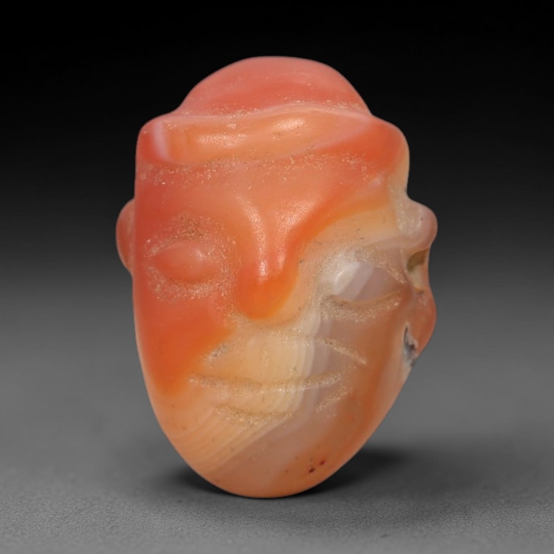 Ancient Agate Face - Near Eastern Amulet Figurine, 37XS: This amulet is carved from banded orange agate, exhibiting natural color gradients and a smooth, polished finish characteristic of ancient Near Eastern glyptic craftsmanship. The form is a stylized hu