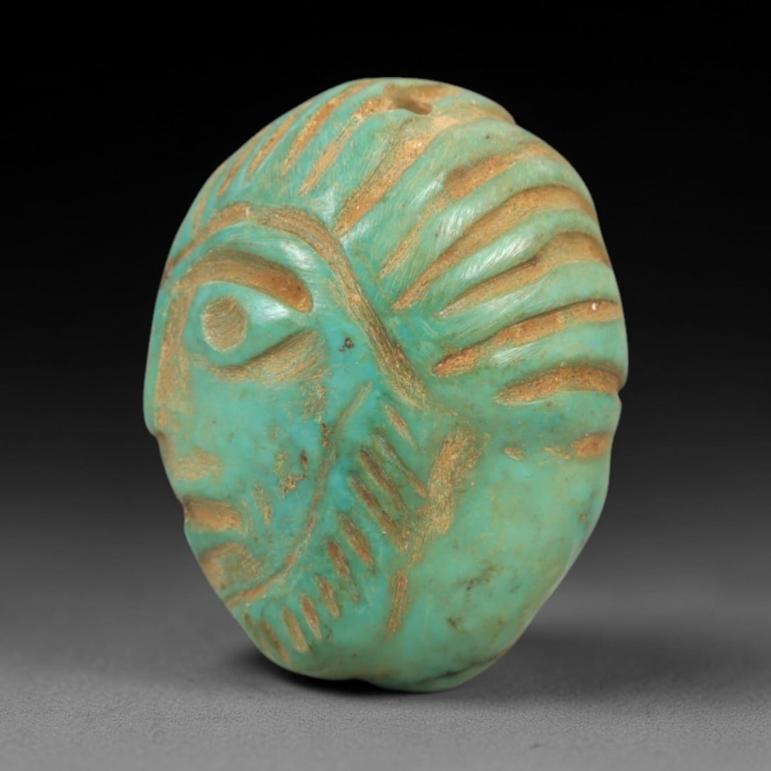 Ancient Turquoise Face - Near Eastern Amulet, 85AK: Carved from polished turquoise, this amulet features a stylized human face in profile, with incised linear details defining the eye, brow, mouth, and striated hair or headdress. A suspension hole at t