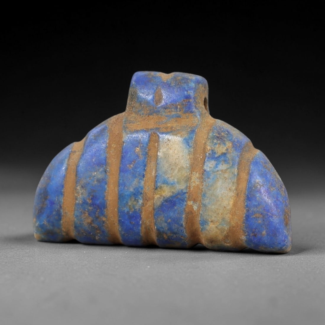 Ancient Lapis Lazuli Amulet - Egyptian Winged Pendant, 16AP: Carved from lapis lazuli, this amulet features a curved, wing-like form with a raised upper lug pierced for suspension, its surface incised with vertical bands that define its segmented structure. The
