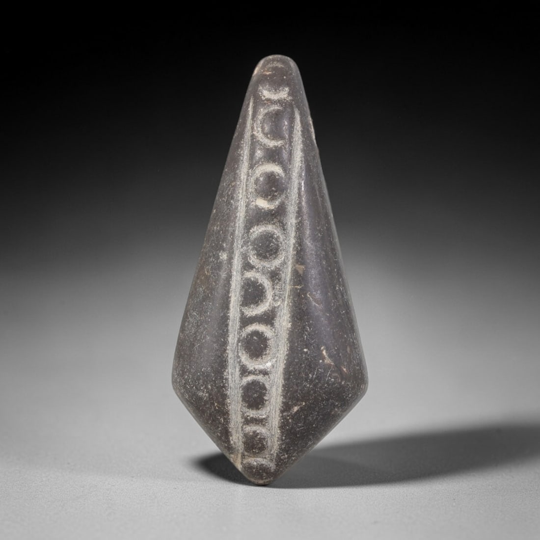 Ancient Stone Amulet - Near Eastern Carved Pendant, 53AR: This artifact is carved from a dense, dark stone with a smooth, weathered patina. The form is elongated and tapering, terminating in a pointed base, with a central vertical incised line flanked by a c