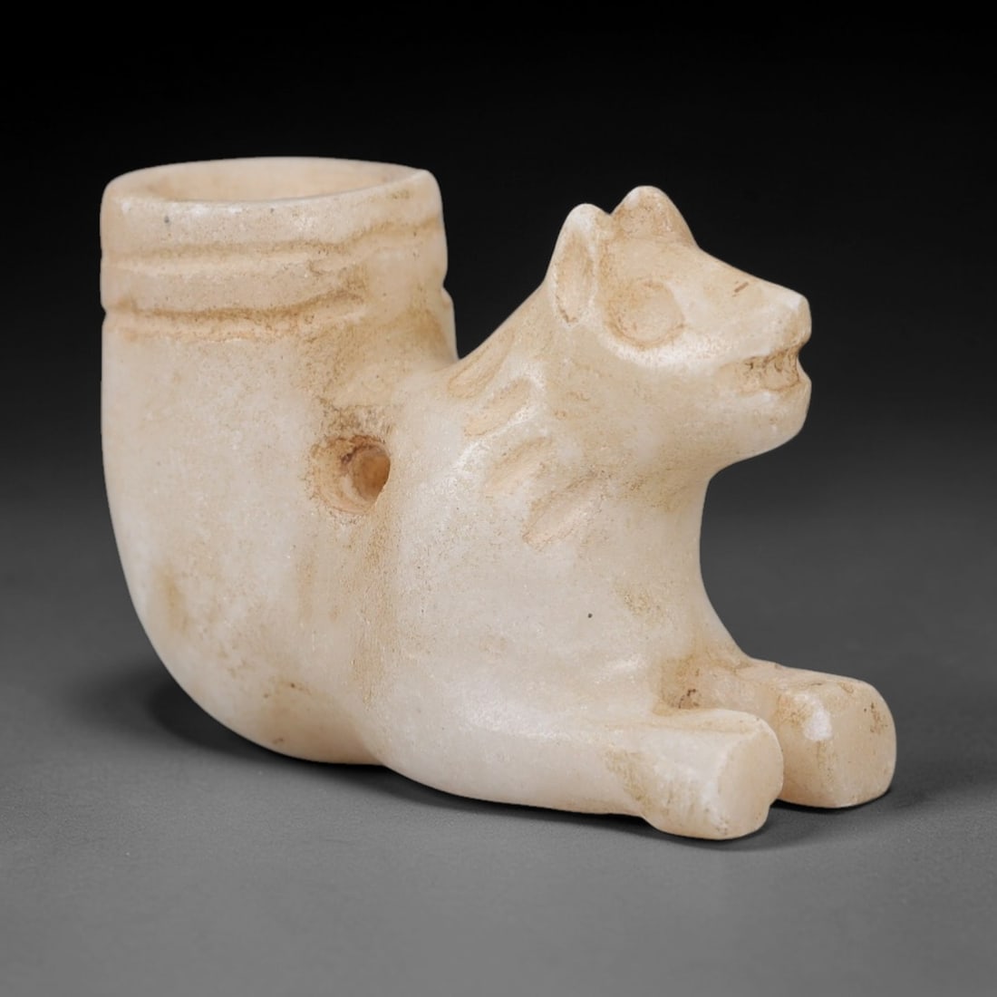 Ancient Marble Rhyton Amulet - Canine Form Figurine, 20AK: Crafted from pale marble, this rhyton-shaped amulet integrates a recumbent canine figure into its curved vessel form. The canine head, detailed with incised linear details defining the mane, rounded e