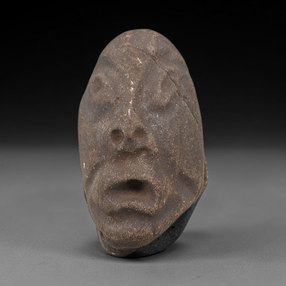 Ancient Stone Maskette - Near Eastern Amulet Figurine, 7FX: Carved from rough, granular stone, this elongated maskette features a stylized human face with recessed eyes, perforated nasal openings, and a large open mouth, creating an expressive, almost vocal ap