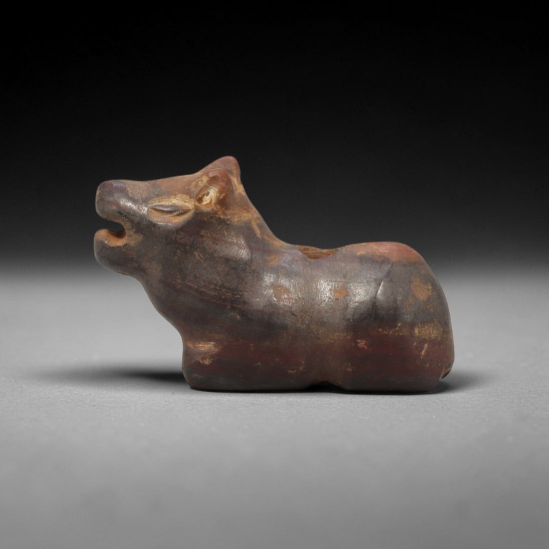 Ancient Stone Bovid Amulet - Near Eastern Figurinev, 8XT: This amulet is carved from a dark reddish-brown stone with subtle tonal banding and a weathered, earthy patina across its surface, executed in a stylized, compact form that emphasizes the recumbent bo