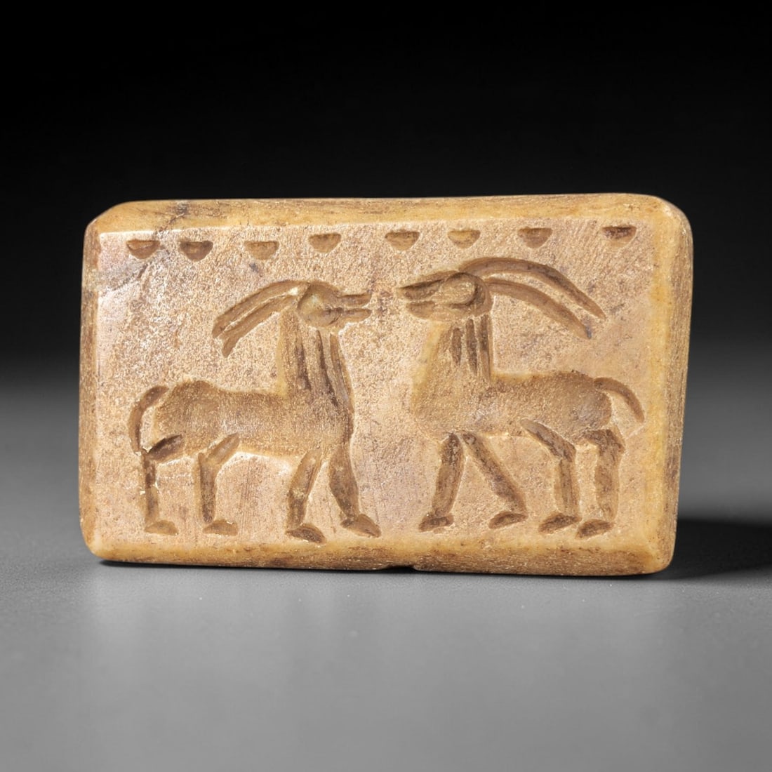 Ancient Steatite Stamp Seal - Near Eastern Ibex Figurine, 55AS: This ancient Near Eastern stamp seal is carved from steatite, shaped as a rectangular plaque with incised decoration depicting two confronting ibex figures in profile, rendered with distinct linear de