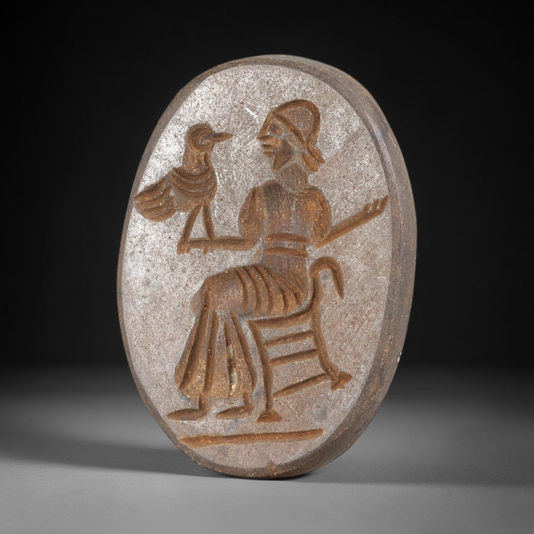 Ancient Stone Stamp Seal - Near Eastern Anthropomorphic Idol, 48AS: This ancient Near Eastern stamp seal is carved from stone, crafted in an oval form featuring a detailed glyptic scene of a seated anthropomorphic figure in profile, holding a bird figure in one outstr