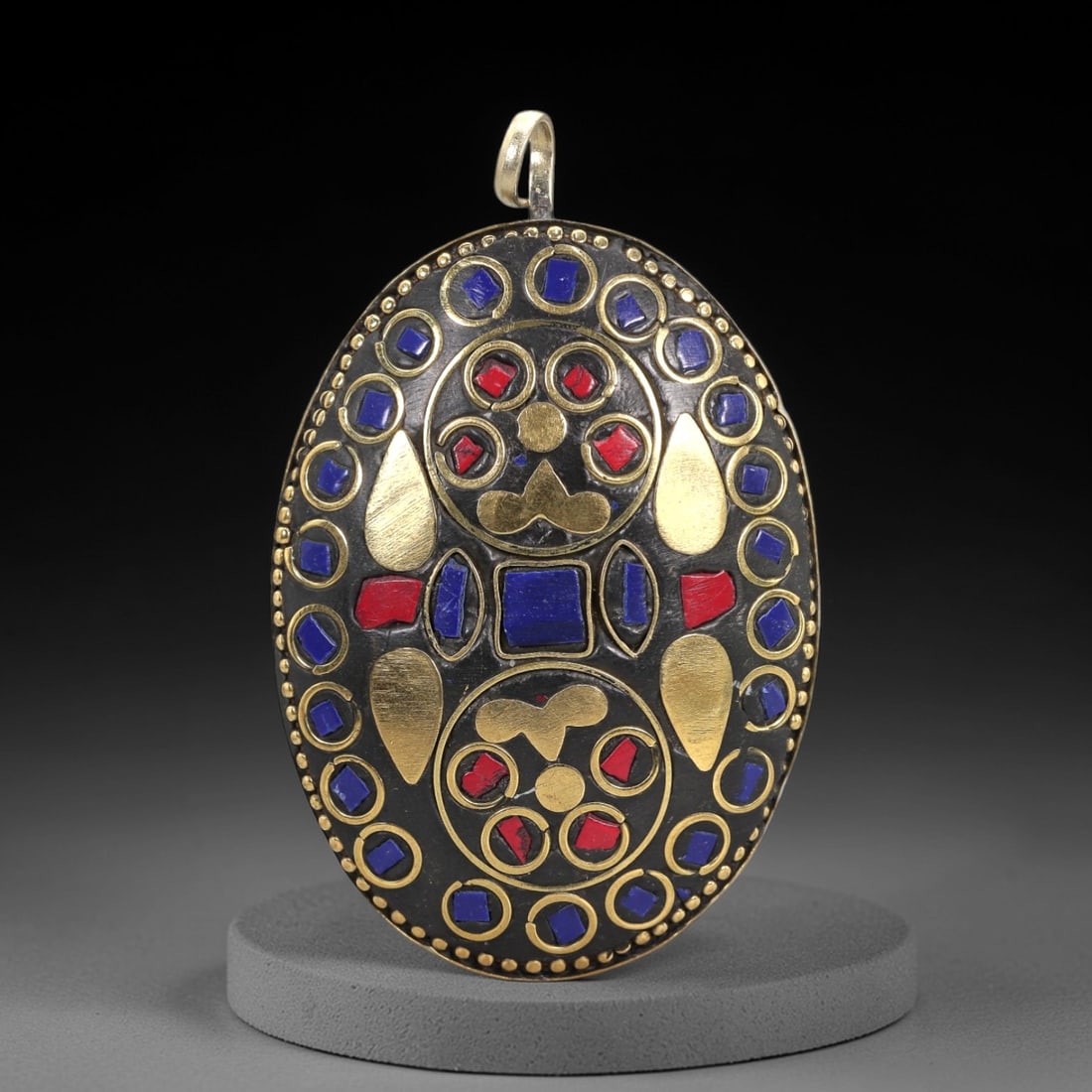 A Tibetan Brass Pendant with Lapis Lazuli and Red Stone Inlay, 20th Century, 97XM: Oval in form, the pendant is crafted from brass with a blackened matrix and features inlays of lapis lazuli, red stone, and applied gilt-brass elements. A single cylindrical brass suspension loop is a