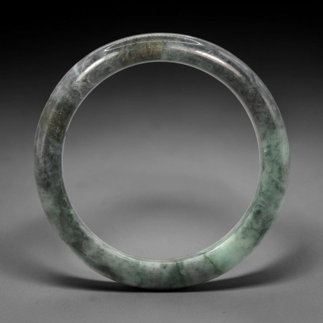 A Chinese Jadeite Jade Bangle Bracelet, 20th Century, 75ZK: Carved from jadeite jade with a variegated palette of pale green and deep greyish-black mottling throughout, the bangle is formed as a continuous circular band with a rounded cross-section. The surfac