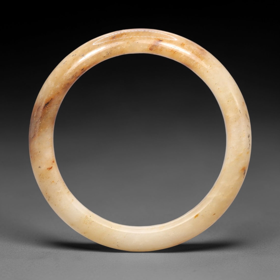A Burmese Jadeite Jade Bangle, 20th Century, 14FS: A Burmese jadeite jade bangle, with a pale creamy-white matrix and scattered russet-brown mineral inclusions. The bangle is of circular form, with a smooth, polished surface revealing the internal cry