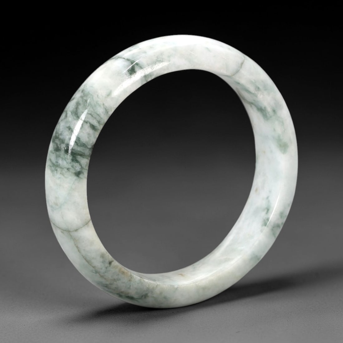A Chinese Jadeite Jade Bangle Bracelet, 20th Century, 81ZK: Carved from jadeite jade with a pale white base and irregular dark green veining throughout, the bangle is formed as a continuous circular band with a rounded cross-section. The surface is smoothly po