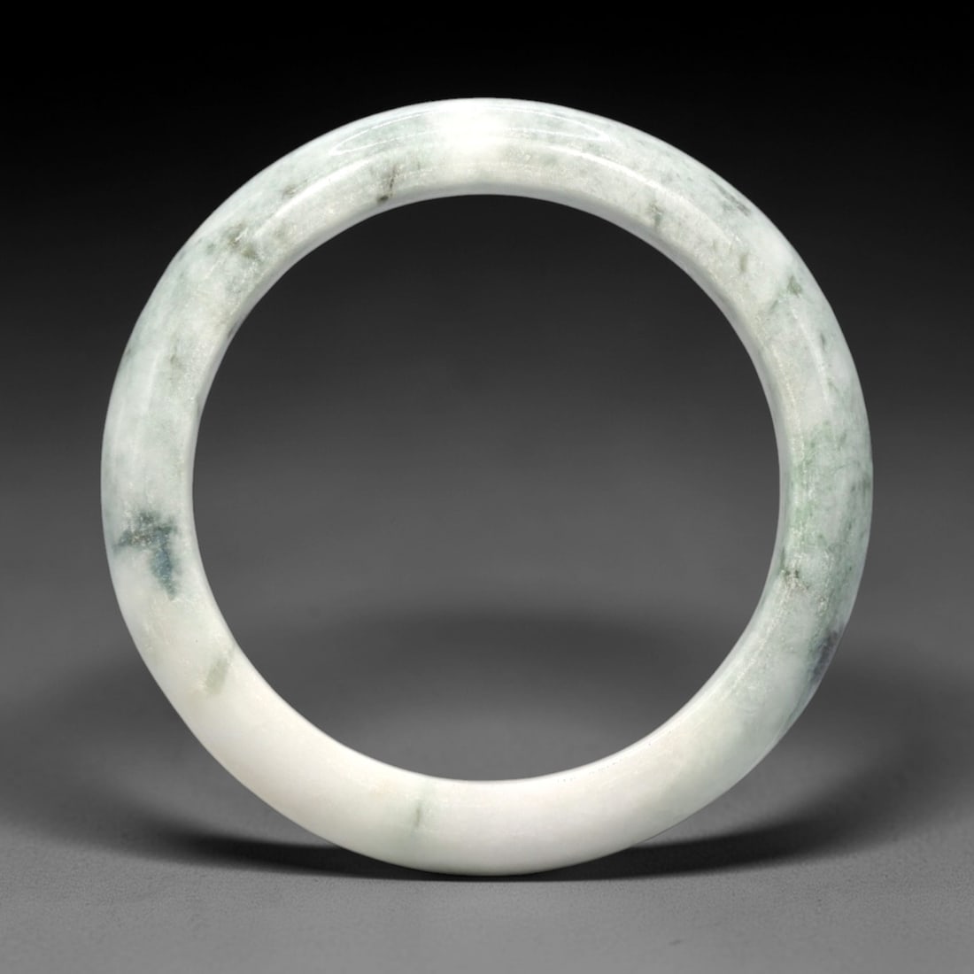 A Burmese Jadeite Jade Bangle, 20th Century, 18FS: A Burmese jadeite jade bangle, with a pale white matrix and scattered dark green mineral inclusions. The bangle is of circular form, with a smooth, polished surface revealing the internal crystalline